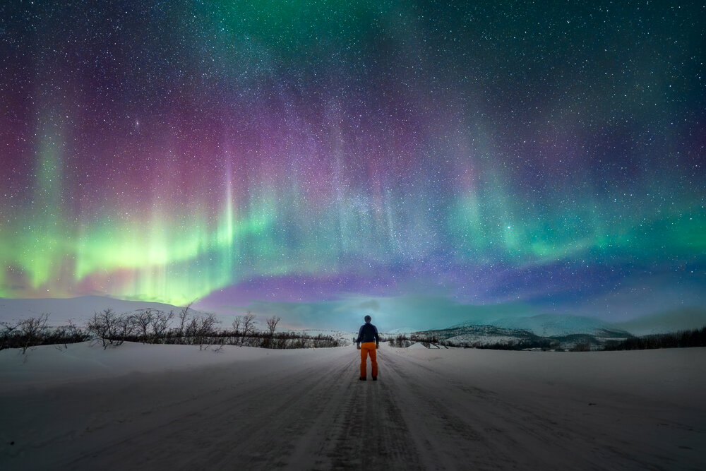 A person stands on a snow-covered road watching a strong northern lights display over the mountains.